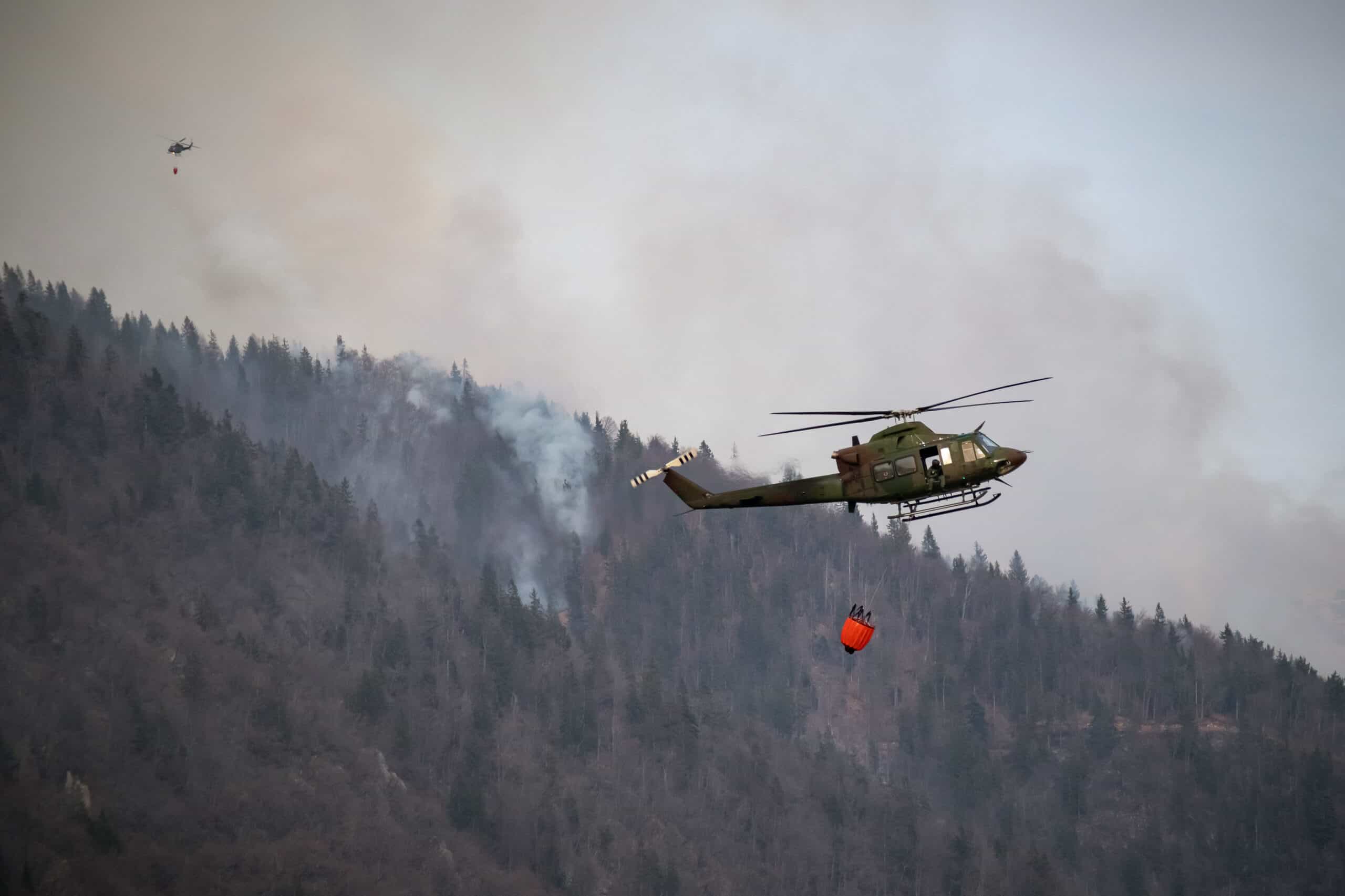 A helicopter dropping water over a forest fire with smoke and flames in the background.