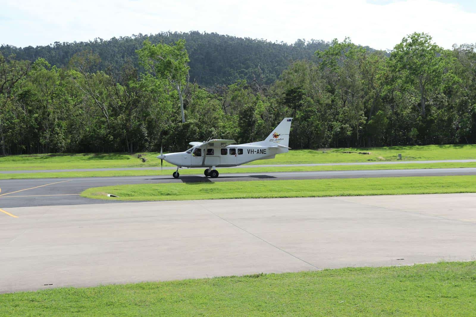 Small propeller plane parked on a runway with lush green trees and hills in the background.