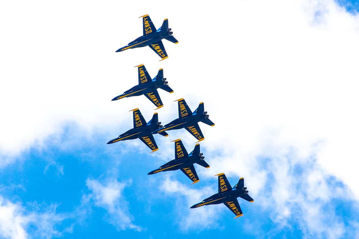 Six US Navy fighter jets flying in tight formation against a blue sky with clouds.